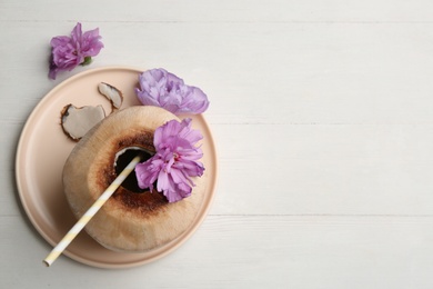 Young peeled coconut with straw and flowers on white wooden table, flat lay. Space for text