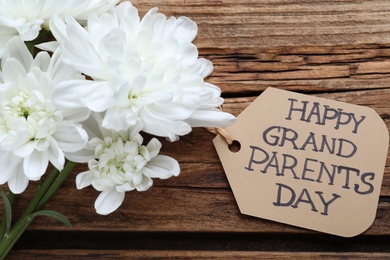 Beautiful white flowers and tag with phrase Happy Grandparents Day on wooden background, flat lay