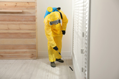 Pest control worker in protective suit spraying insecticide on floor at home