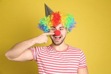 Emotional young man with party hat and clown wig on yellow background. April fool's day
