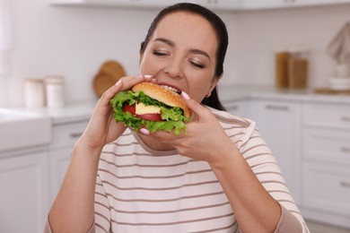 Overweight woman eating burger in kitchen at home