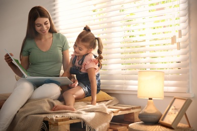 Young woman and her daughter reading book near window at home