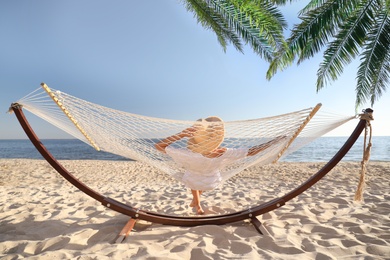 Woman relaxing in hammock under green palm leaves on sunlit beach