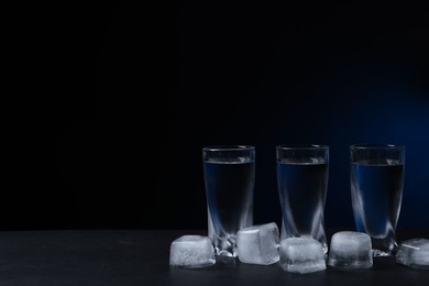 Shot glasses of vodka with ice cubes on black table against dark background. Space for text