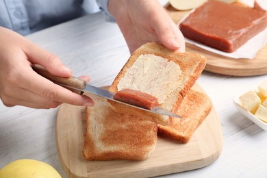Woman spreading delicious quince paste on toast bread at white wooden table, closeup