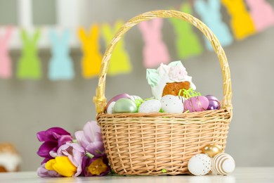 Photo of Wicker basket with painted eggs and delicious Easter cake near tulips on table indoors