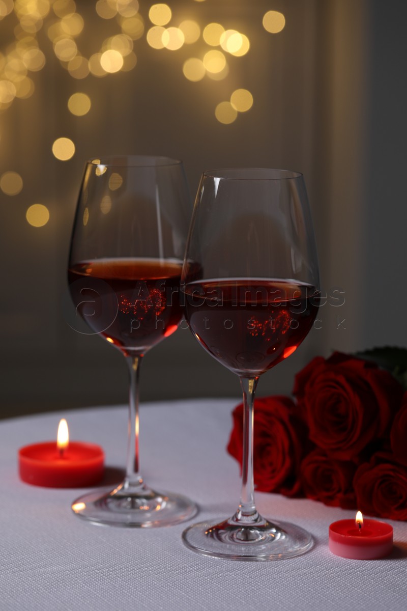 Photo of Glasses of red wine, rose flowers and burning candles on white table against blurred lights. Romantic atmosphere