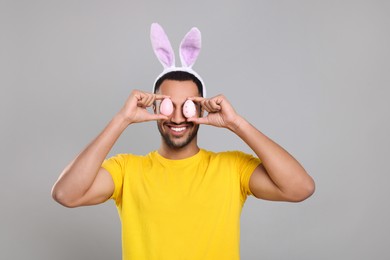 Photo of Happy African American man in bunny ears headband covering eyes with Easter eggs on gray background