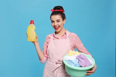 Housewife holding bottle of cleaning product and basin with clothes on light blue background