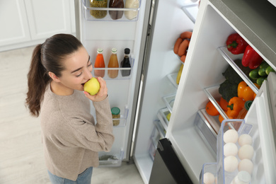 Young woman eating apple near open refrigerator indoors, above view