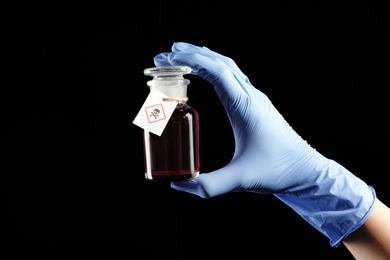 Woman in gloves holding glass bottle of poison with warning sign on black background, closeup
