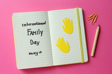 Notebook with text International Family Day May 15, pencil and paper clips on pink background, flat lay