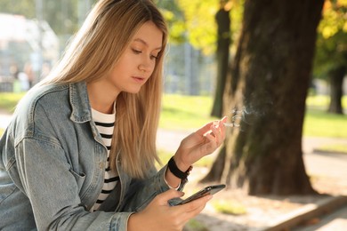 Photo of Young woman smoking cigarette while using smartphone outdoors