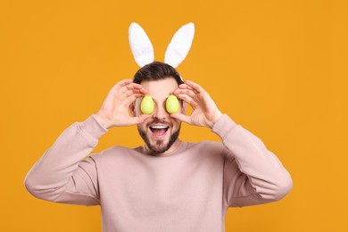 Photo of Happy man in bunny ears headband holding painted Easter eggs near his eyes on orange background
