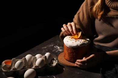 Young woman decorating traditional Easter cake at table against black background, closeup. Space for text