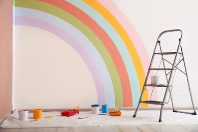 Different decorator's tools and ladder near wall with painted rainbow indoors