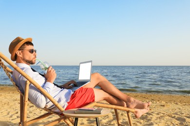 Photo of Man with laptop and drink resting on beach. Business trip