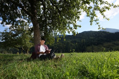 Handsome man reading book under tree on meadow in mountains