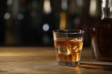 Photo of Glass of whiskey with ice on wooden table in bar, space for text