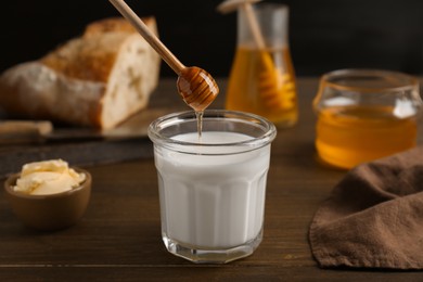 Photo of Pouring honey from dipper into glass with milk near butter on wooden table