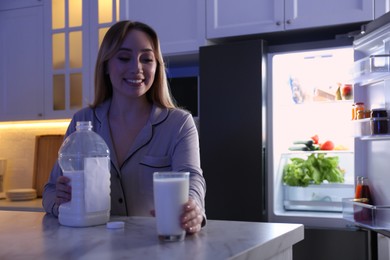 Young woman holding glass and gallon bottle of milk on white marble table in kitchen at night. Space for text