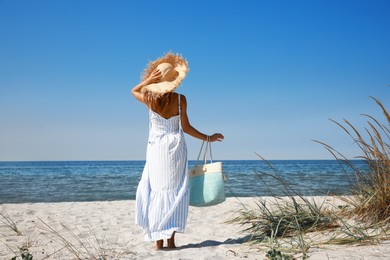 Woman with beach bag and straw hat on sand near sea, back view