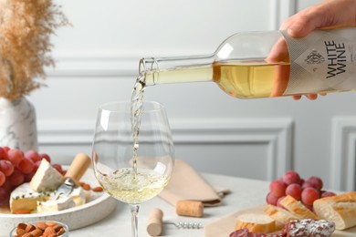 Woman pouring white wine from bottle into glass at table with snacks, closeup