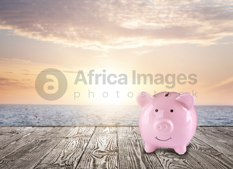 Saving money for summer vacation. Piggy bank on wooden surface near sea at sunset, space for text
