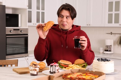 Overweight man with glass of cold drink and tasty croissant at table in kitchen