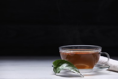 Photo of Aromatic hot tea in glass cup and leaf on white table against black background. Space for text
