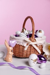 Photo of Wicker basket with festively decorated Easter eggs and bunny on white marble table against pink background