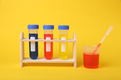 Photo of Beaker and test tubes with colorful liquids in wooden stand on yellow background. Kids chemical experiment set