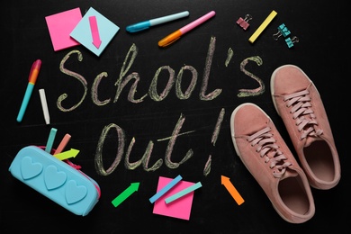 Shoes, stationery and text SCHOOL'S OUT written on blackboard, flat lay. Summer holidays