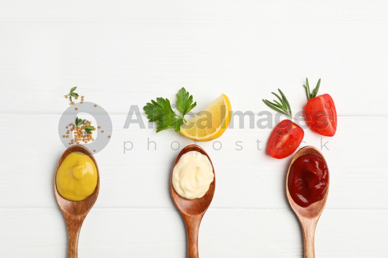 Ketchup, mayonnaise, mustard in spoons and ingredients on white wooden table, flat lay