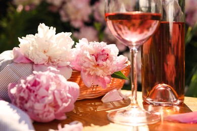 Bottle and glass of rose wine near beautiful peonies on wooden table in garden, closeup
