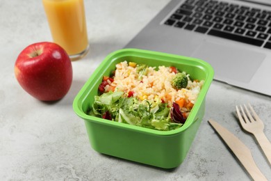 Photo of Bowl of tasty food, laptop, apple and cutlery on light grey table, closeup. Business lunch