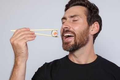 Happy man eating sushi roll with chopsticks on light grey background, closeup