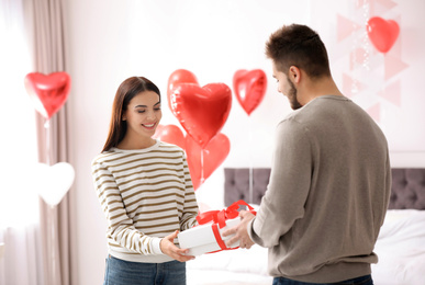 Young man presenting gift to his girlfriend in bedroom decorated with heart shaped balloons. Valentine's day celebration