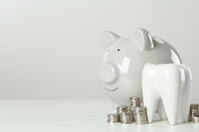 Ceramic model of tooth, piggy bank and coins on white table, space for text. Expensive treatment
