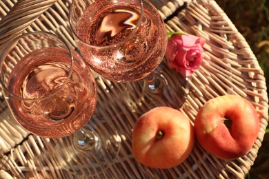 Photo of Glasses of delicious rose wine, flower and peaches on wicker basket outside, above view. Space for text