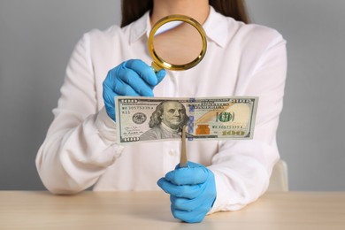 Photo of Expert authenticating 100 dollar banknote with magnifying glass at table on light grey background, closeup. Fake money concept