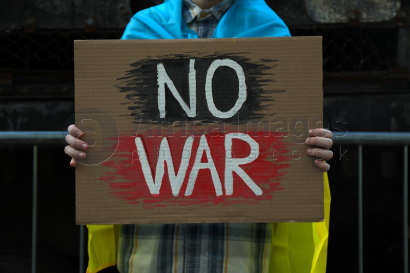 Woman wrapped in Ukrainian flag holding poster with words No War on street, closeup
