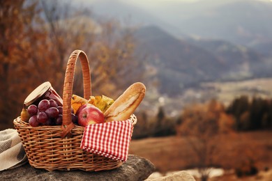 Wicker picnic basket with different products on rock in mountains. Space for text