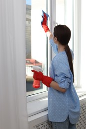 Young woman cleaning window glass with rag and detergent at home, back view