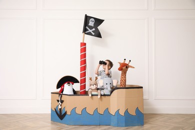 Cute little boy playing with binoculars in pirate cardboard ship near white wall indoors