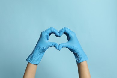 Doctor in medical gloves making heart with hands on light blue background, closeup
