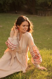 Beautiful woman in embroidered dress sitting on green grass outdoors. Ukrainian national clothes