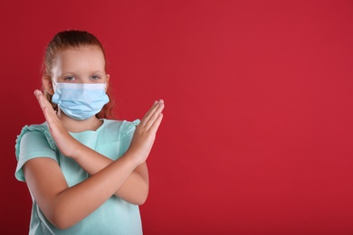 Little girl in protective mask showing stop gesture on red background, space for text. Prevent spreading of coronavirus