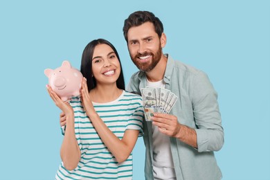 Happy couple with money and ceramic piggy bank on light blue background