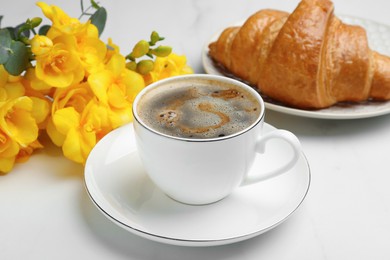 Cup of aromatic coffee, beautiful yellow freesias and croissant on white table. Good morning